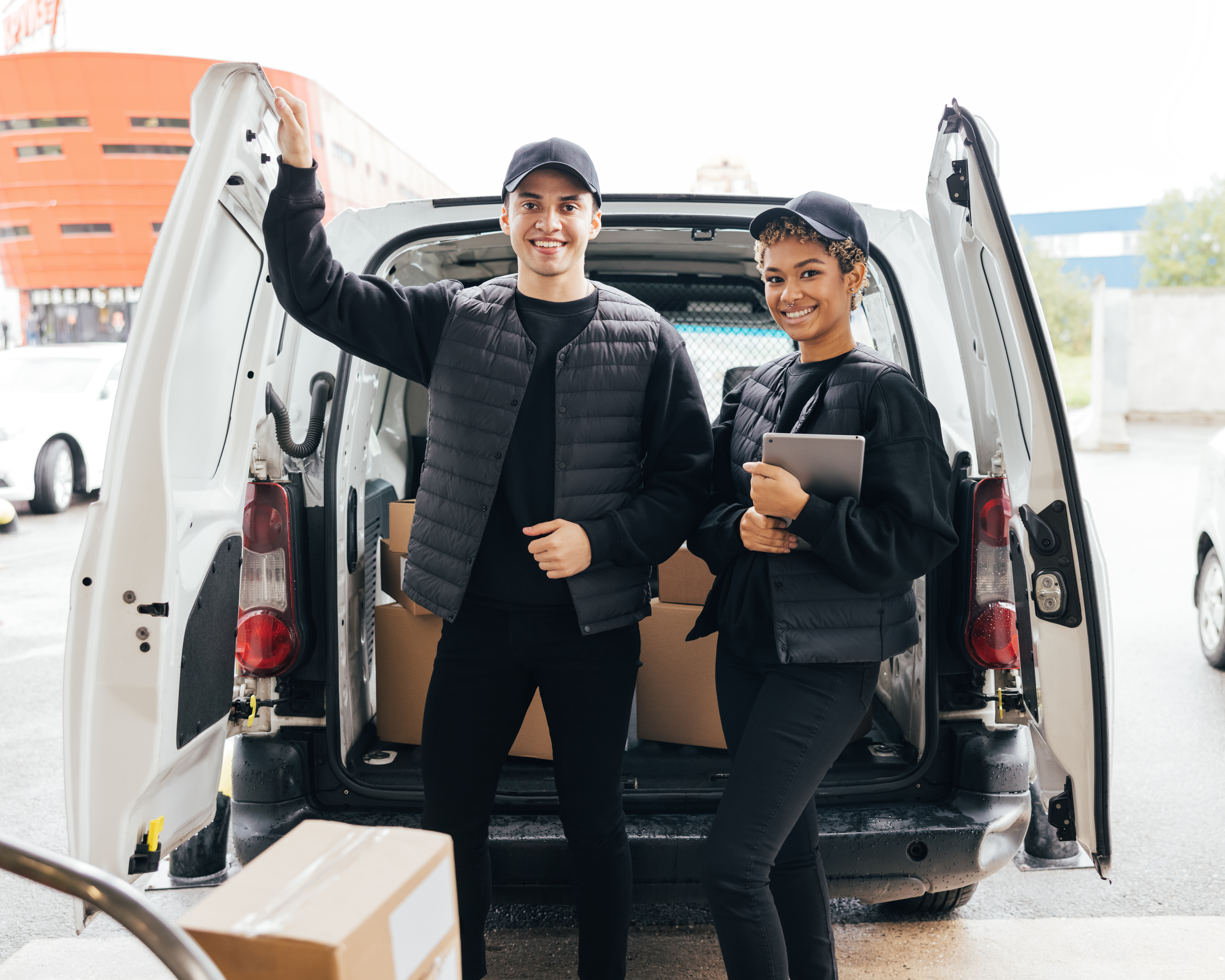 Portrait of two young couriers in uniform standing at van trunk and looking at camera